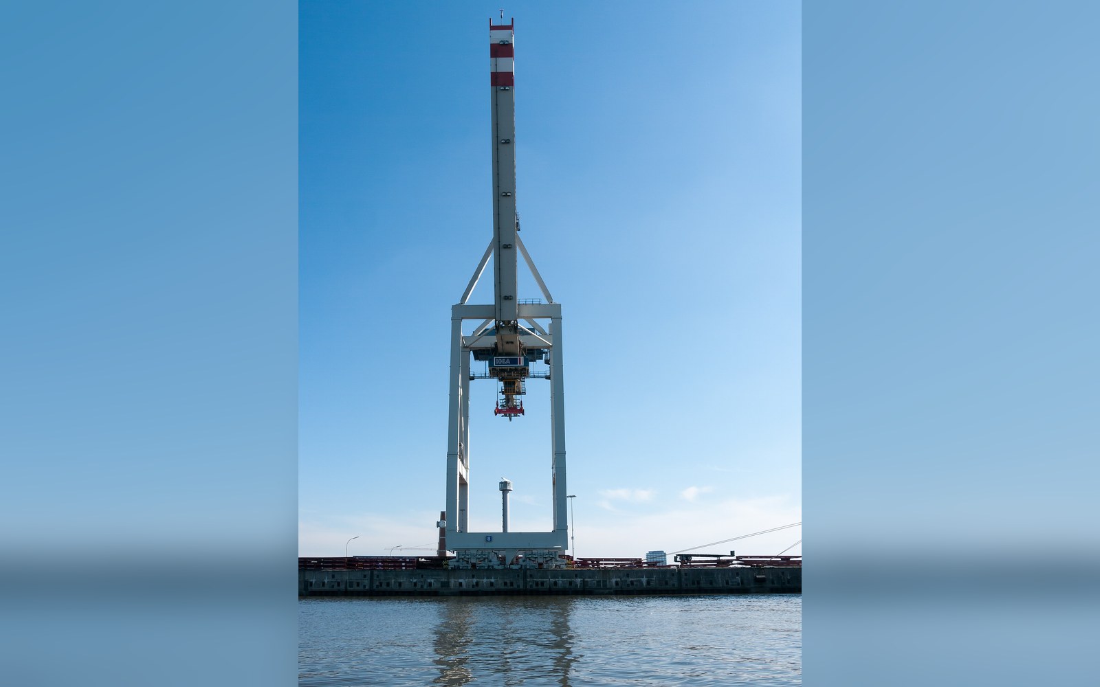 Tall gantry crane with raised boom stands at a waterside dock under clear sky