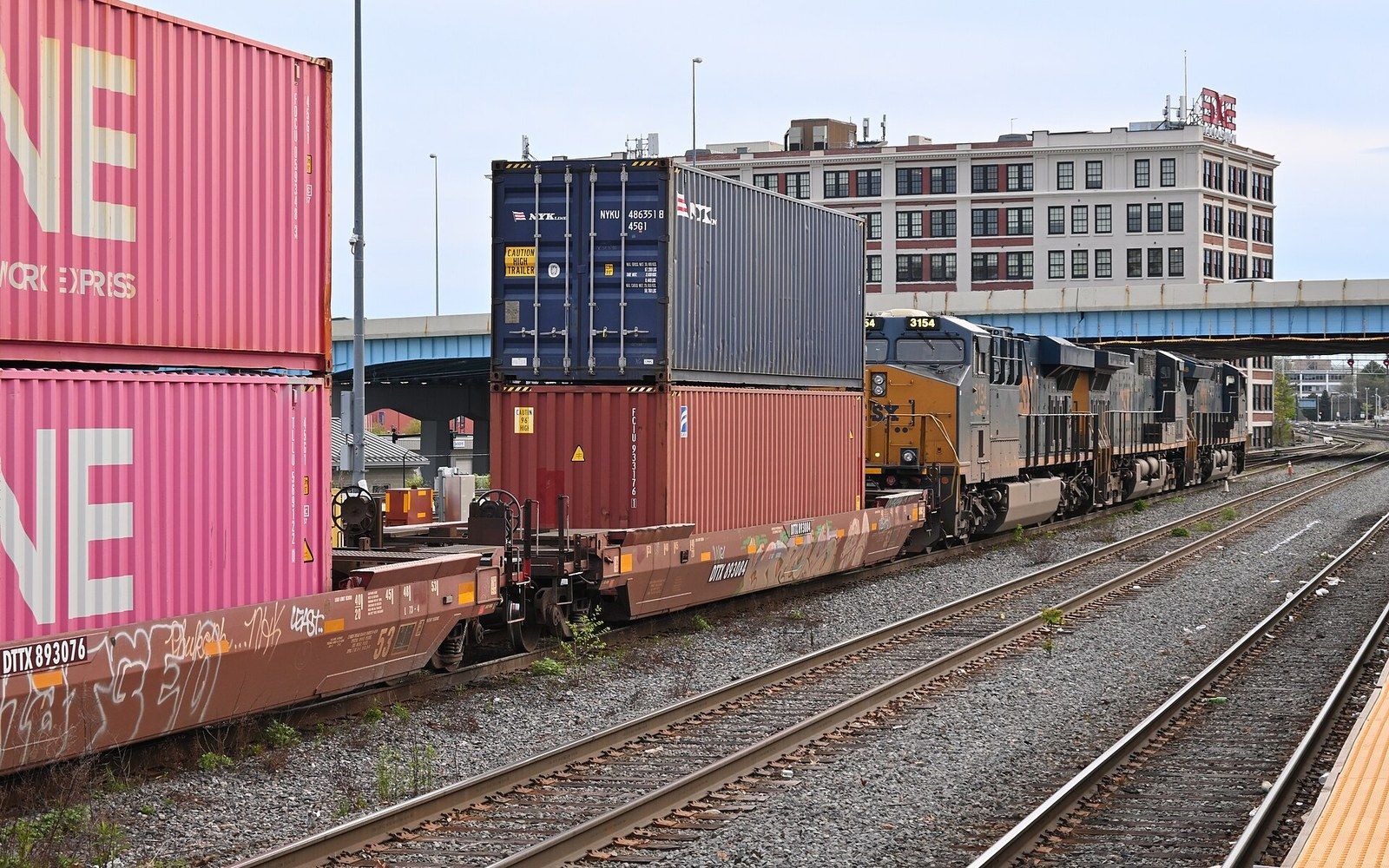 Stacked shipping containers ride double-stack rail cars behind a locomotive in a rail yard